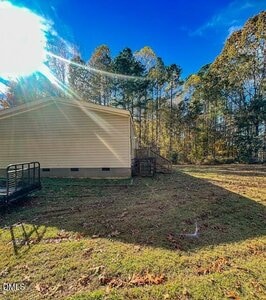 A beige single-story house with a small set of wooden steps at the side, surrounded by a grassy yard and trees under a clear blue sky with sunlight streaming in.