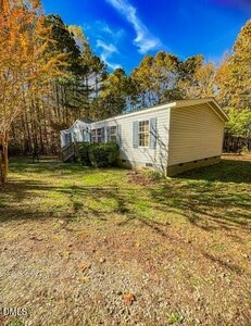A small, light-colored manufactured home with a front porch sits on a grassy lot surrounded by trees under a clear blue sky. Sunlight casts shadows across the yard.