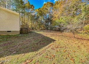 A grassy backyard area with scattered leaves, bordered by woods and tall trees, is shown next to the side of a light-colored house with a small wooden staircase. The sky is clear and blue.