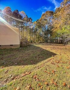 A grassy yard with scattered leaves next to the side of a beige house. Trees with autumn foliage stand in the background under a clear blue sky. The house casts a shadow on part of the yard.