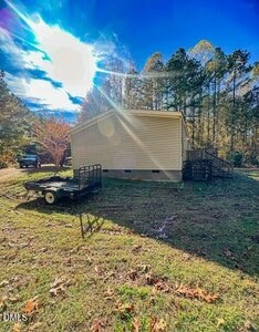 A single-story house with beige siding is shown from the side, with a small trailer parked on the grassy yard. Trees surround the property, and the bright sun is visible in a blue sky.