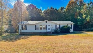 A single-story white house with a gable roof sits on a large grassy lawn at 7537 Sam Young Road in Oxford. The home features several windows, a small porch with steps, and is surrounded by tall trees under a clear blue sky.