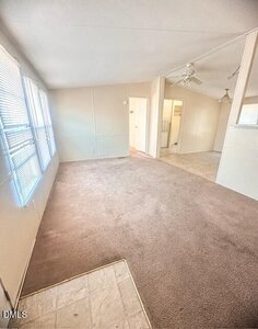 A room with beige carpet, light-colored walls, and large windows along one side. There is a ceiling fan, a tiled entryway, and an open doorway leading to another space.