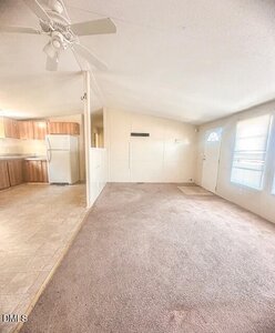 A bright, empty living area with carpeted flooring next to a kitchen with tile flooring, wood cabinets, a refrigerator, and a ceiling fan. Sunlight comes through two windows near the front door.