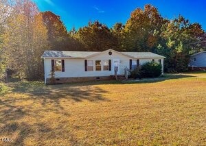 A single-story white house with dark shutters sits on a large grassy lot, bordered by tall trees under a clear blue sky. The house has a small front porch and minimal landscaping.