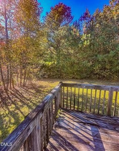 A wooden deck with railing overlooks a grassy yard bordered by dense trees under a clear blue sky. Sunlight casts shadows across the deck and lawn.