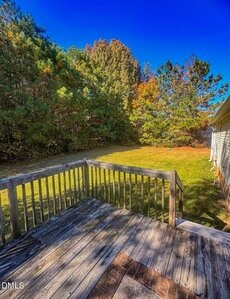 Wooden deck with railing overlooking a grassy backyard bordered by trees. The sky is clear and bright blue, and some of the trees display fall colors. Part of a light-colored house is visible on the right.