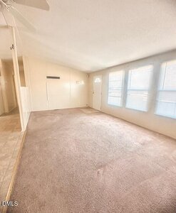 A bright, empty living room with beige carpet, three large windows with blinds, a white front door, and an adjoining tiled hallway. The walls are painted a light color and the ceiling is slightly vaulted.