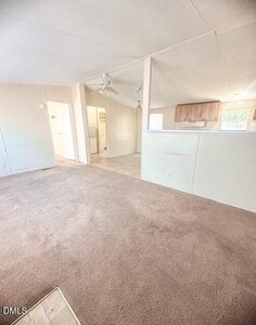 A room with beige carpet, white walls, and a partial divider separating the carpeted area from a linoleum-floored space. Ceiling fan, doorway to a bathroom, and a small kitchen with wooden cabinets are visible.