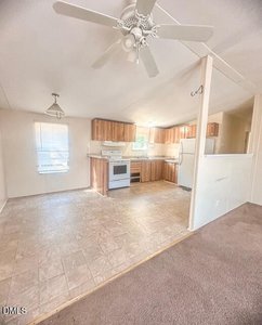 A kitchen with wood cabinets, white appliances, and a ceiling fan is shown next to a dining area with a window. The floor is linoleum, and brown carpet marks the transition to another room.