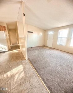 A small, unfurnished living space with beige walls, a carpeted area, tile flooring near the kitchen, and a white door with windows letting in natural light. A refrigerator is visible at the end of the hallway.