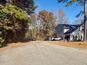 A quiet suburban street bordered by trees with two parked vehicles in the driveway of a two-story house on the right side. Fallen leaves are scattered across the road and lawns. The sky is clear and blue.