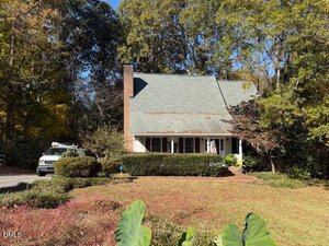 A two-story house with a gray roof, brick chimney, and white siding is surrounded by trees and shrubs. A white van is parked in the driveway to the left. The yard has trimmed grass and mature landscaping.