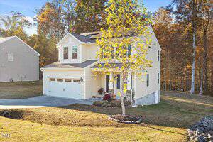 A two-story beige house with white trim and a two-car garage sits on a sloped lawn. A small tree is in the front yard, and autumn-colored trees are in the background. The driveway curves up to the garage.