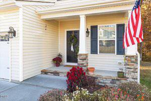 A beige house with a front porch features a black door with a wreath, potted plants on the steps, stone accents, outdoor lighting, and an American flag displayed on a white column.