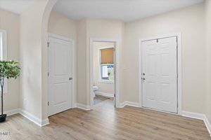 A clean entryway with light wood flooring, beige walls, two white doors, an arched doorway, a small window, and a potted plant in the corner. A bathroom is visible through the open doorway.