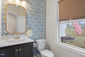 A bathroom with a blue patterned wallpaper, a white sink with a gold-framed mirror and gold fixtures, a white toilet, and a window with a beige shade partially open, showing an American flag outside.