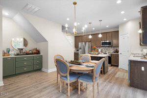 A modern kitchen and dining area with wood floors, round dining table with four chairs, green sideboard with drinks under stairs, brown cabinets, stainless steel appliances, and pendant lighting.