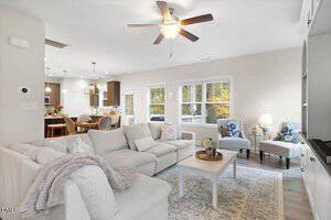 A bright living room with a white sectional sofa, a patterned rug, a coffee table with flowers, two armchairs, and a ceiling fan. Large windows bring in natural light, and a dining area is visible in the background.