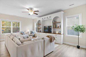 A bright living room with a white sectional sofa, a TV on a built-in entertainment center with shelves, a coffee table, two armchairs, large windows, a ceiling fan, and a potted plant.