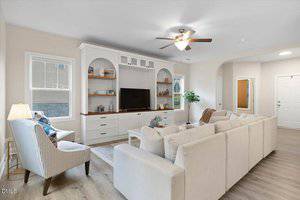 A living room with a large white sectional sofa, a striped armchair, a white entertainment center with shelves and cabinets, a TV, decorative items, wood flooring, and a ceiling fan with lights.