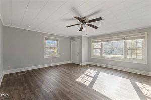 Empty room with light gray walls, white ceiling, dark wood floor, ceiling fan with light, large windows letting in sunlight, and a white door; space appears bright and clean.