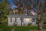 A small, single-story house at 312 11th Street in Butner features gray siding, black shutters, and a dark roof. Surrounded by grass, trees, and bushes, sunlight brightens the front beneath a sky of blue and purple hues.
