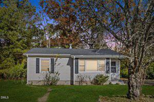 A small, single-story house at 312 11th Street in Butner features gray siding, black shutters, and a dark roof. Surrounded by grass, trees, and bushes, sunlight brightens the front beneath a sky of blue and purple hues.