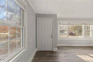 A small, empty room with gray walls, white trim, and dark wood flooring. A white front door is centered, and large windows on two walls let in natural light and show trees and grass outside.
