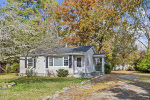 A small, single-story gray house with white trim sits on a grassy lot surrounded by trees with autumn leaves. A gravel driveway curves in front of the house, and there is a small covered entry porch.