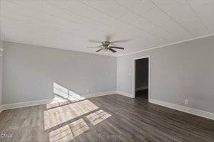 An empty room with light gray walls, white trim, wood-look flooring, a ceiling fan with dark blades, and natural light streaming in through a window. An open doorway leads to another room.