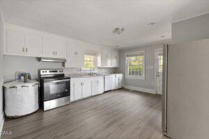 A kitchen with white cabinets, stainless steel appliances, a roll of paper towels, and wood-look flooring. There is a window above the sink and a glass door leading outside. The room is bright and uncluttered.