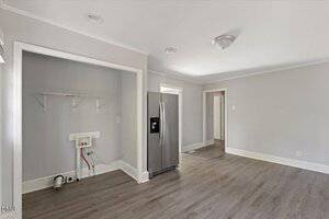 A room with light gray walls and wood flooring, featuring a stainless steel refrigerator, an empty laundry nook with shelves and hookups, and a ceiling light fixture. There are doorways leading to other rooms.