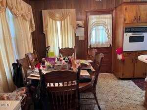 A dining area with wood-paneled walls, a round wooden table cluttered with various items, six wooden chairs, curtained windows and door, and a built-in oven in a wood cabinet. The floor is covered in patterned linoleum.