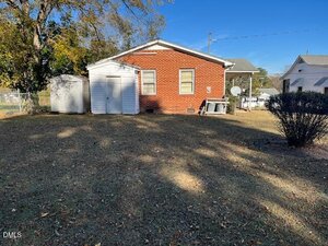 A small red brick house with two white storage sheds in a grassy, partly shaded yard. There are two windows on the back wall, a bush to the right, and a chain-link fence on the left. Other homes are visible in the background.