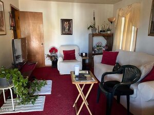 A living room with red carpet, white furniture, red and white cushions, a TV on a stand, a small wooden tray table, black plastic chair, houseplants, and shelves with decor against cream-colored walls and curtains.