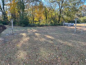 A grassy backyard with scattered fallen leaves, two wooden clothesline poles, and surrounding trees with autumn foliage. A fenced area and more trees are visible in the background.