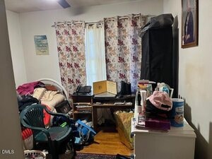 A small cluttered room with a single bed, a plastic chair, a desk with electronics and papers, laundry baskets, and floral curtains covering a window. Various items are scattered around, including clothing and cleaning supplies.