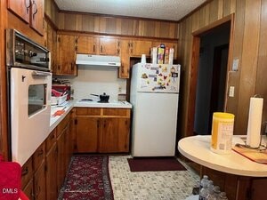 A small kitchen with wood-paneled walls, wood cabinets, a white fridge with magnets, a white oven, a stove, a counter with cleaning supplies, and a patterned rug on a tiled floor. Boxes and paper towels are on top of the fridge.