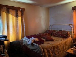 A dimly lit bedroom with yellow curtains, a metal bed frame, a beige comforter, multiple pillows, and some clothes on the bed. Light from the window creates a warm glow on the walls and furniture.