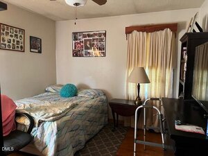 A small bedroom with a single bed, a teal round pillow, a walker, a desk with a TV, a lamp on a folding table, light curtains covering the window, and family photos on the walls.