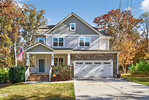 A two-story suburban house with gray siding, stone accents, and a covered front porch. An American flag hangs by the entrance. The house has a double garage and is surrounded by trees with autumn foliage.