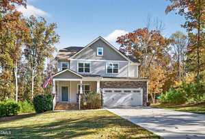 A two-story house with gray siding, stone accents, and a covered front porch. An American flag is displayed near the entrance. A driveway leads to a double garage. Trees with autumn foliage surround the property.