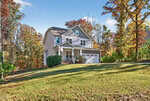 A two-story house at 1318 Sourwood Drive in Wake Forest, with gray siding and white trim, features a covered front porch, an American flag, and a two-car garage, set on a grassy lawn among autumn trees under a blue sky.