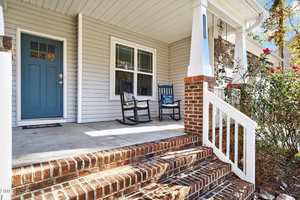 A front porch with brick steps, two black rocking chairs, a small side table, and a light blue door. White railings and beige siding are visible, with some flowering bushes by the stairs.