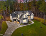 A gray, two-story suburban house with a three-car garage is surrounded by a green lawn and bordered by tall trees. The driveway curves toward the house, and exterior lights are on at dusk.