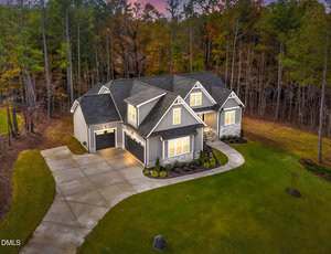 A gray, two-story suburban house with a three-car garage is surrounded by a green lawn and bordered by tall trees. The driveway curves toward the house, and exterior lights are on at dusk.