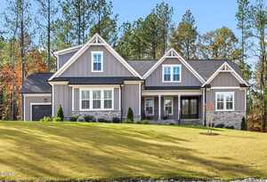 A modern suburban house with gray siding, white trim, and a front porch sits on a neatly manicured lawn at 3925 Cedar Knolls Drive in Youngsville. Tall pine trees and autumn foliage are visible in the background.