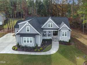 A gray, single-story house with white trim and a black roof sits on a landscaped lot with a driveway, front porch, and a curved sidewalk; trees and grass surround the property.