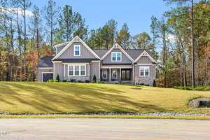 A modern single-story house with gray siding and stone accents sits on a large, sloped lawn with young landscaping, surrounded by tall trees under a clear blue sky.
